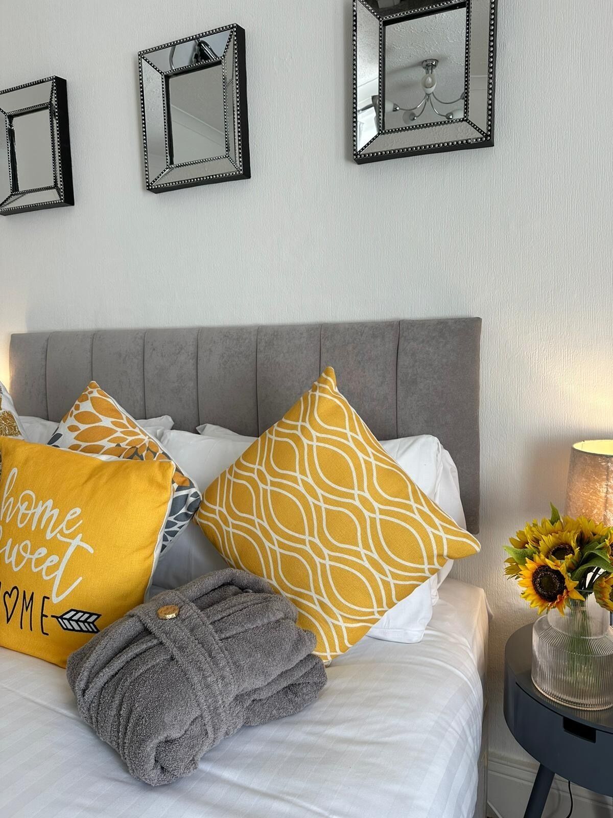 Un lit bien fait, avec une tête de lit grise, plusieurs oreillers aux couleurs jaune et blanche, une serviette grise et un vase rempli de tournesols, posé sur une table de chevet.