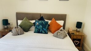 A neatly made bed with a striped white bedspread, flanked by two nightstands on either side. Each nightstand holds a lamp and a small box, possibly for toiletries. The room features a simple, clean design with a plain wall and a wooden headboard above the bed. - Abbey House Bed and Breakfast (Penrith)