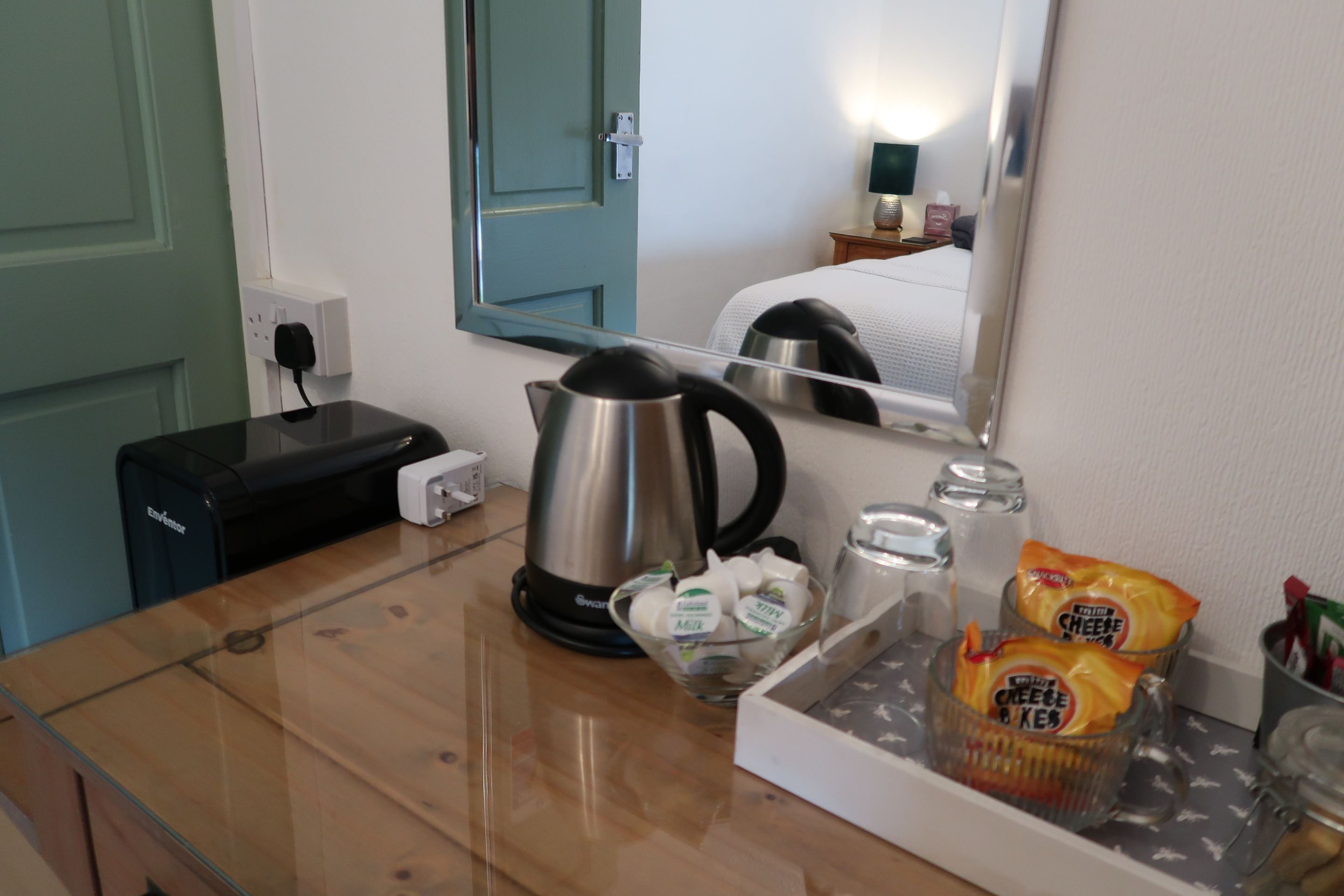 Habitación de hotel con una mesa de madera, un hervidor, un vaso de leche, una bolsa de galletas de queso y un espejo que refleja la habitación.