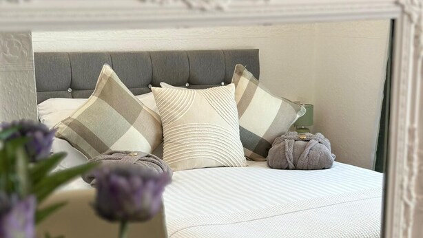A bedroom with a bed, pillows, a bedside table with a lamp, a remote control, and a vase with flowers. - Abbey House Bed and Breakfast (Penrith)