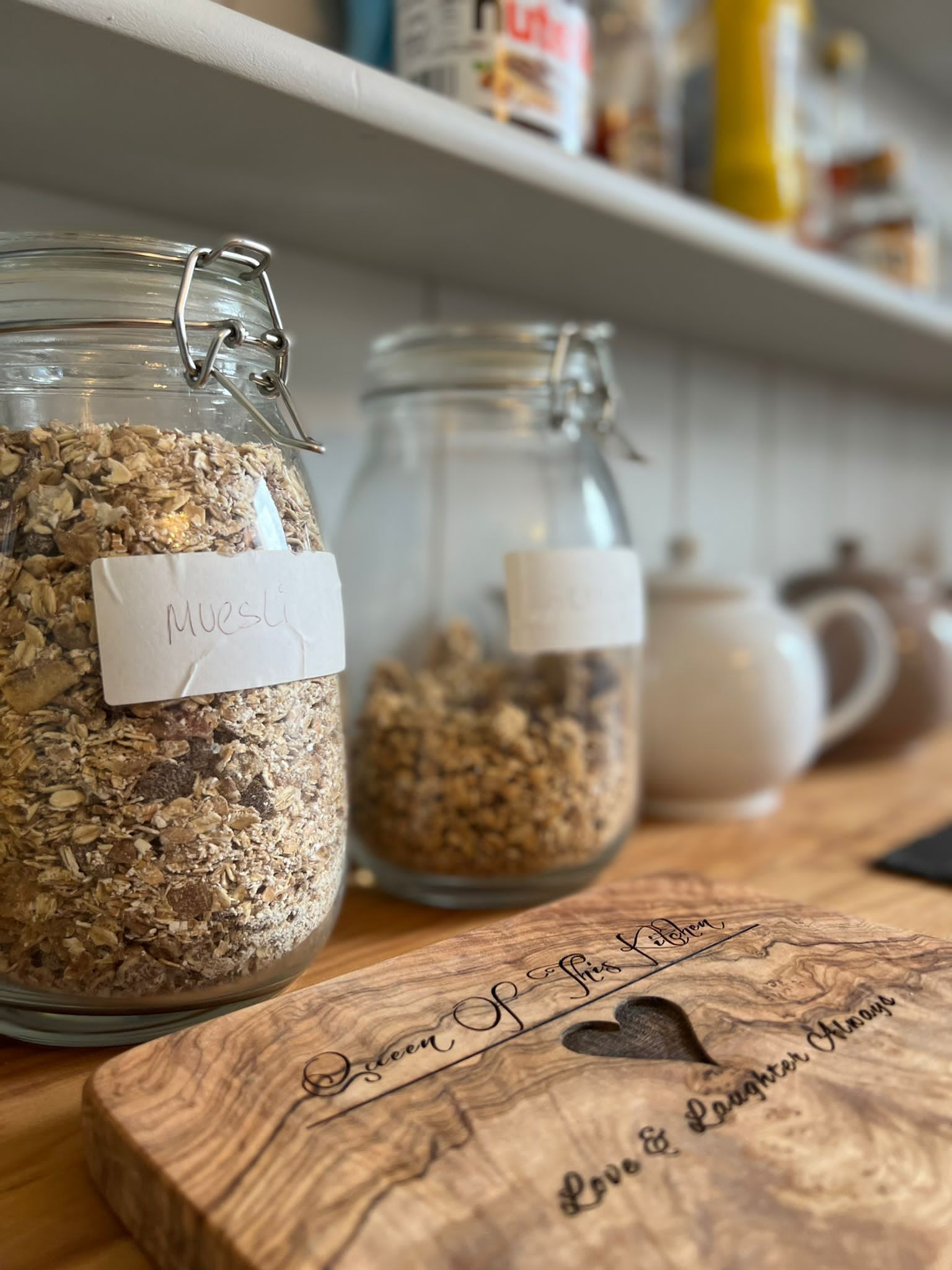Three glass jars with labels on a wooden surface, a white mug, and a shelf with various items in the background.