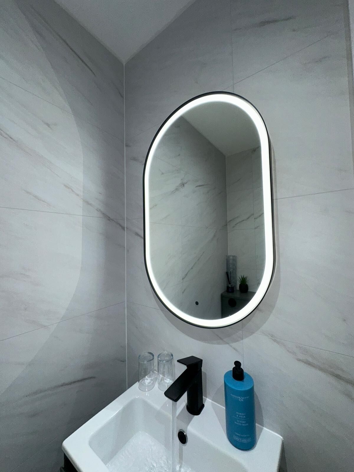 Un baño con un lavamanos blanco, un grifo negro, una botella azul y un espejo con marco iluminado.