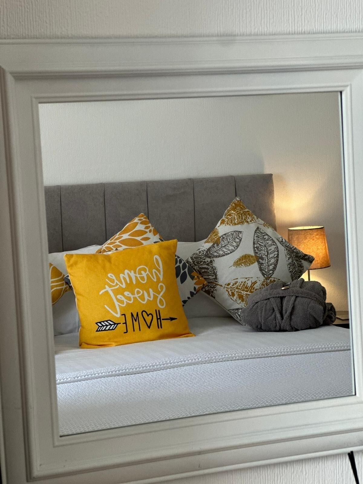 Een bed met een wit hoofdeinde, twee decoratieve kussens en een bedlamp.
