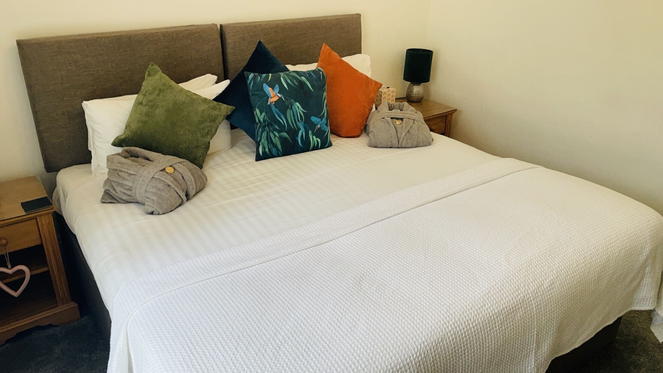 A neatly made bed with a white bedspread, multiple pillows in various colors, and a small bedside table with a lamp and a box.