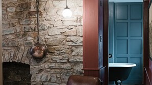 A room with a stone wall, a brown leather chair, a hanging light fixture, and a view of a bathroom with a blue door. - Bridge House (Tiverton)
