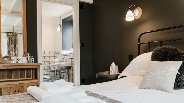 A neatly made bed with white linens, a wooden nightstand with a lamp, and a bathroom visible through an open door. - Bridge House (Tiverton)