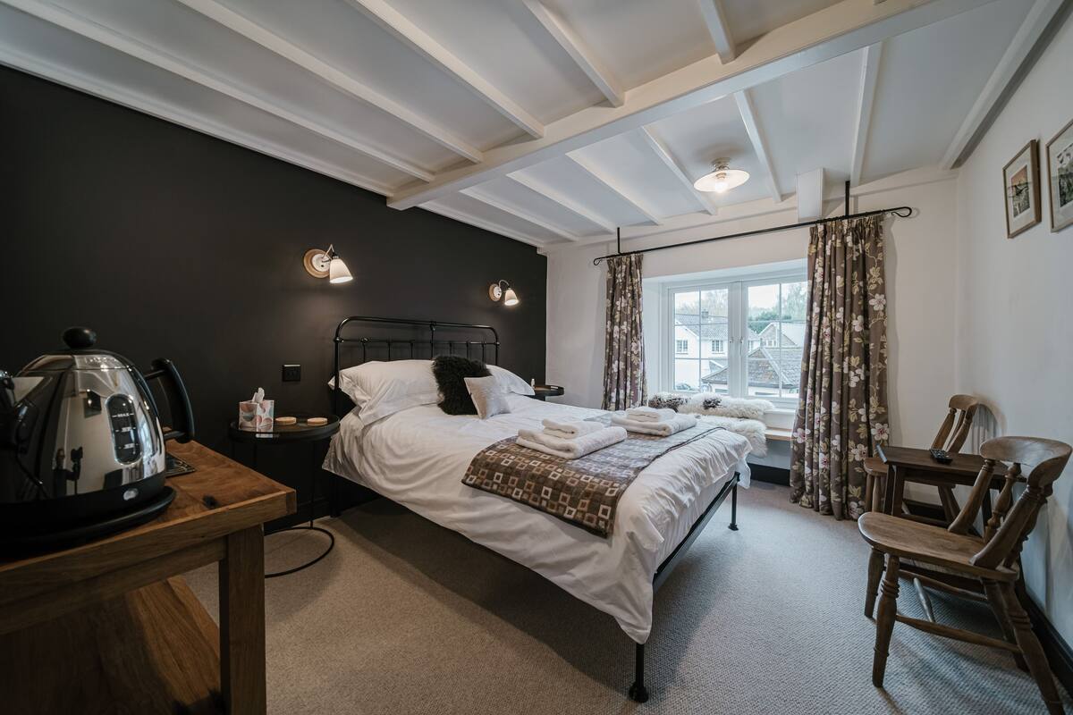 A hotel room with a bed, a wooden table with a kettle, a chair, and a window with curtains.