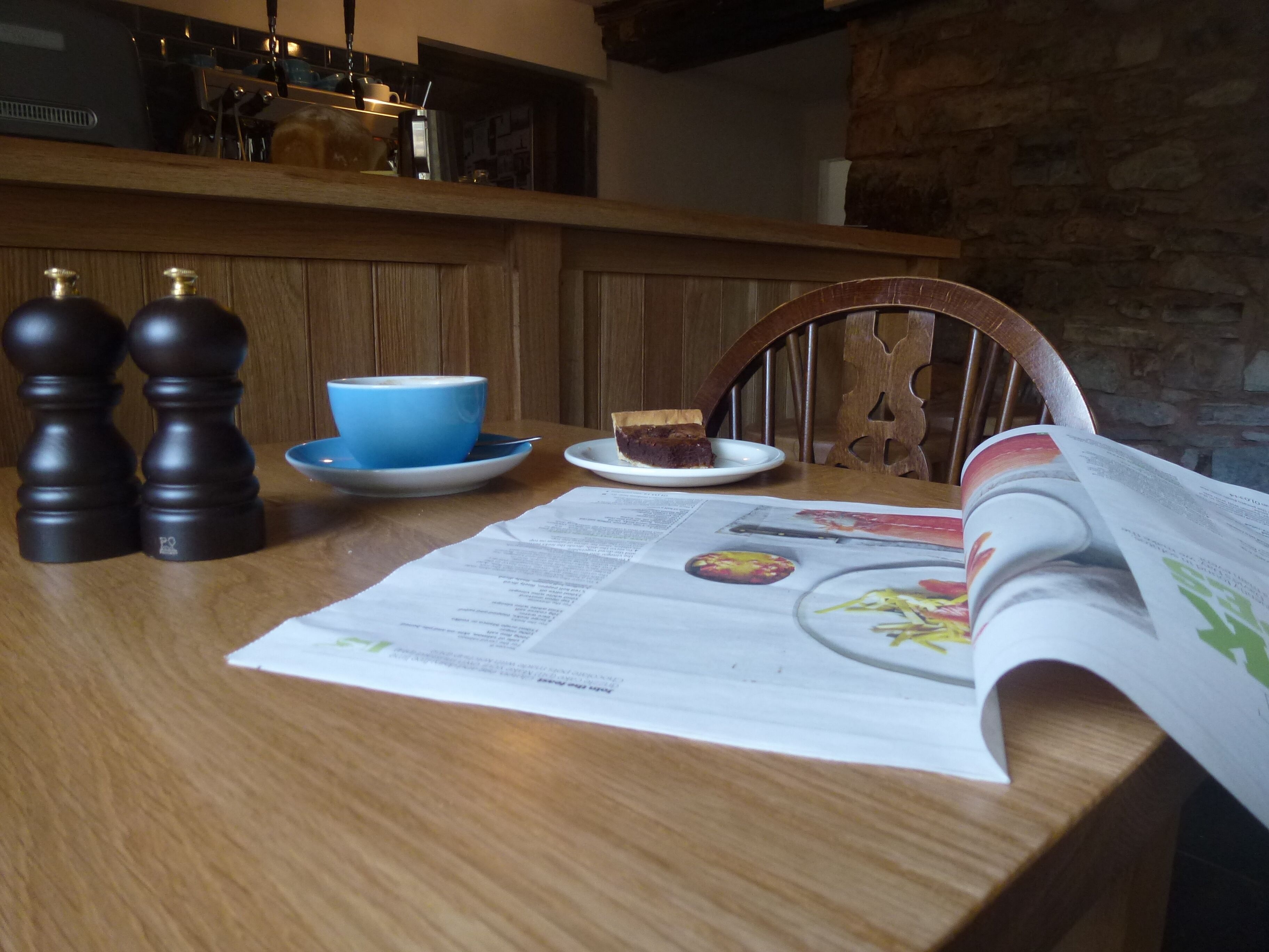 A wooden table with a menu, a blue cup, a plate with a slice of cake, and two salt shakers.