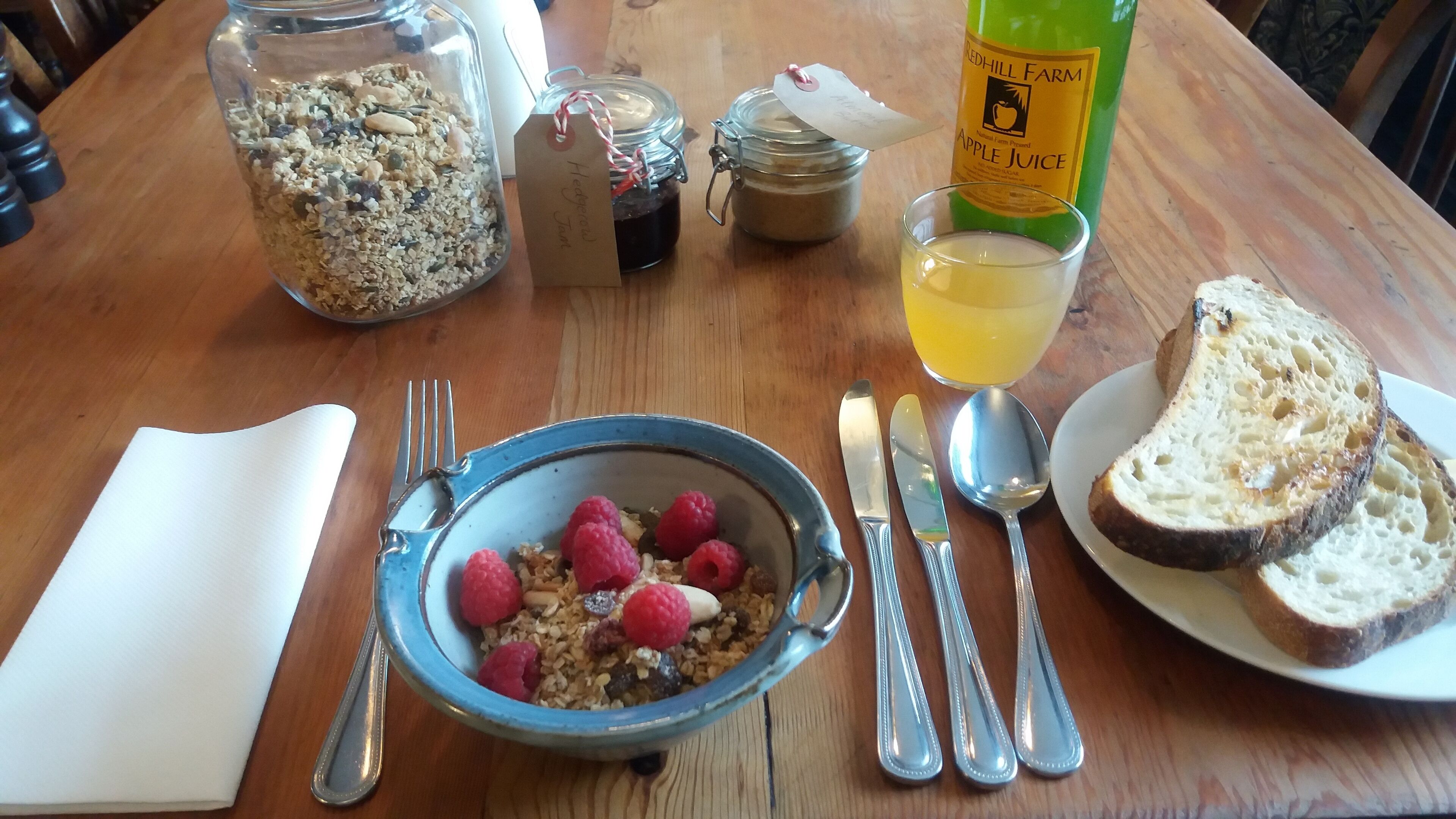 A breakfast setting with a bowl of cereal and raspberries, a glass of juice, and bread on a wooden table.