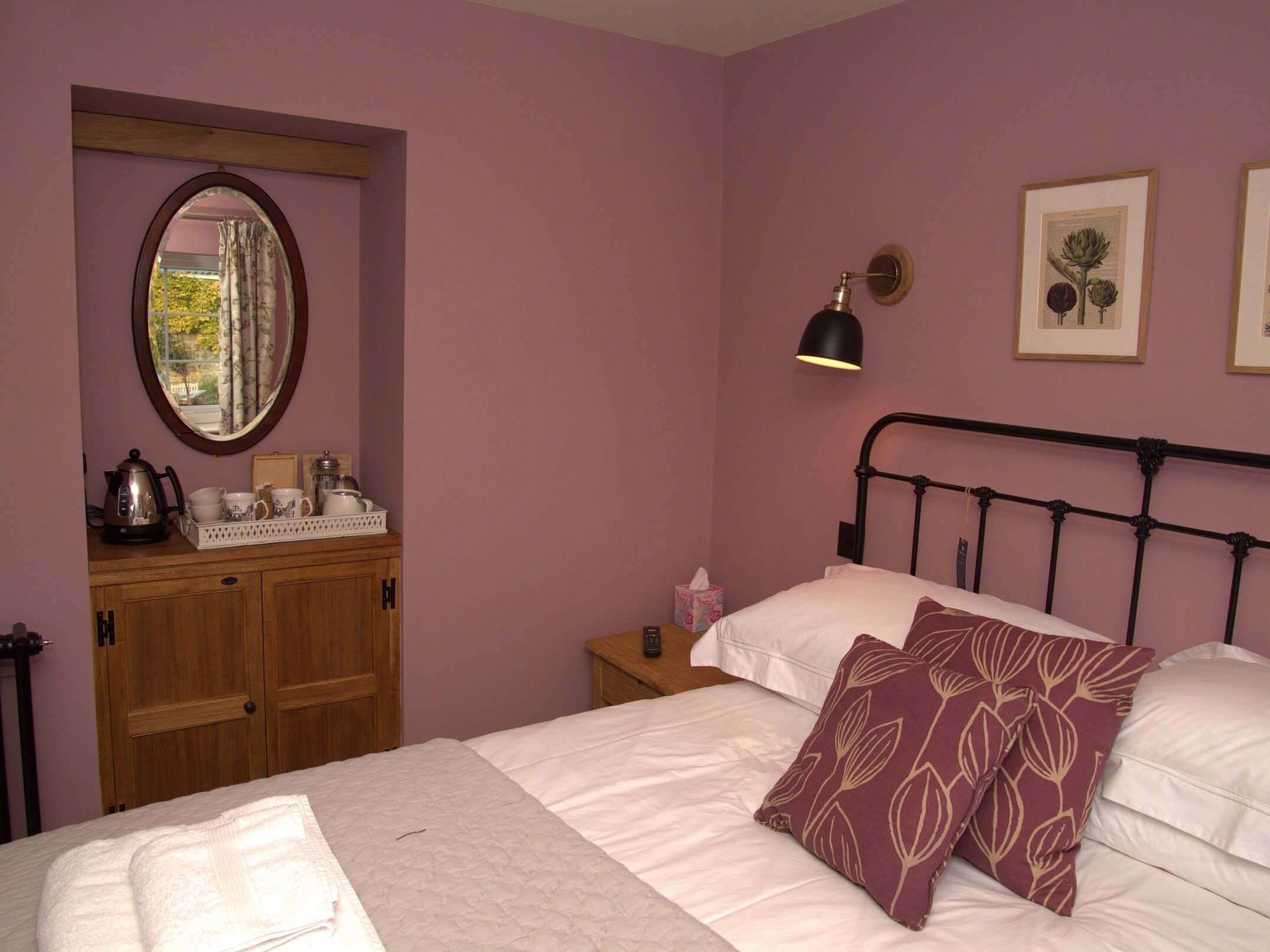 A bedroom with a bed, a nightstand, a mirror, a window with curtains, and framed botanical prints on the wall.
