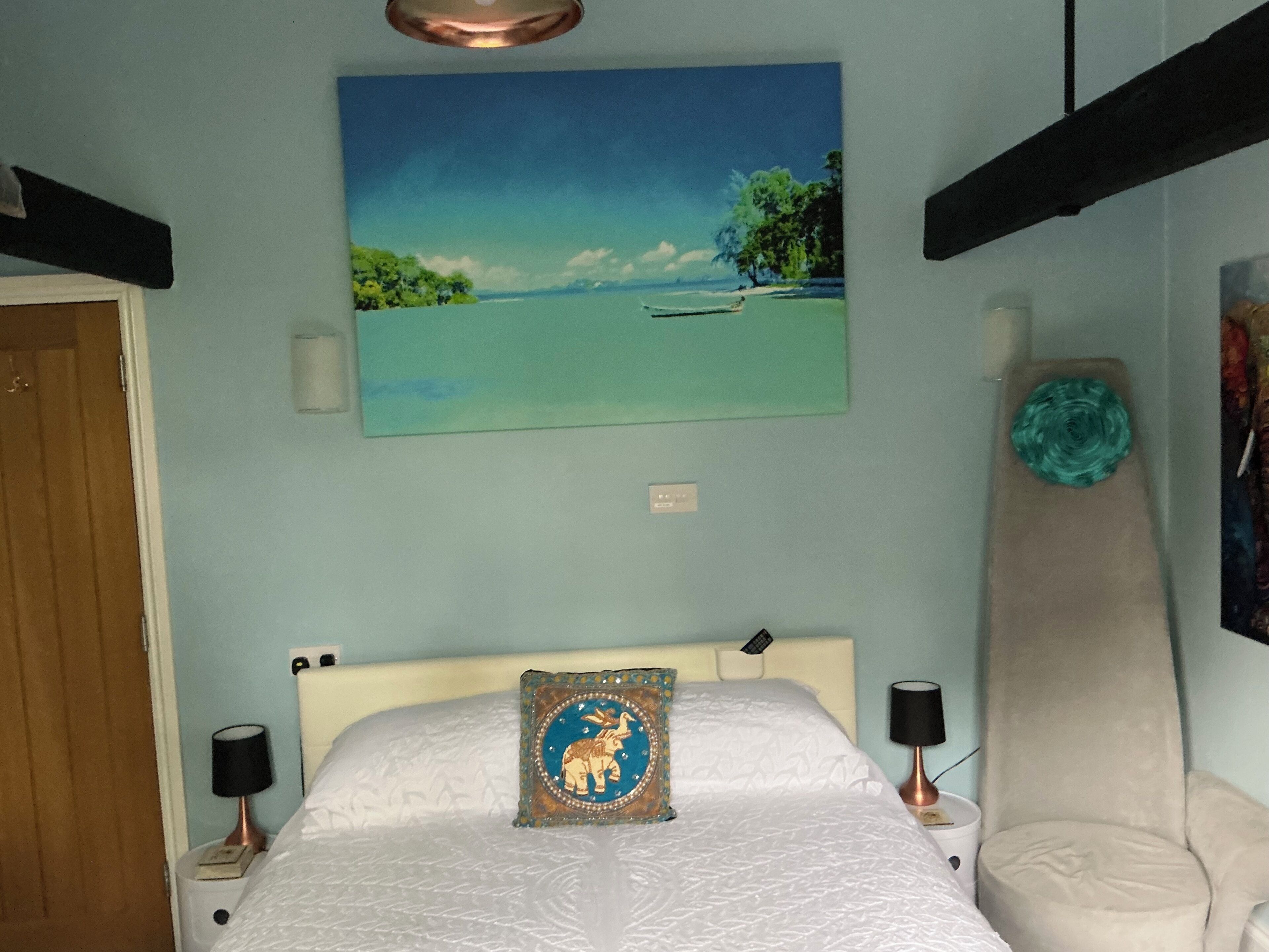 A bedroom with a bed, a painting of a beach scene, a nightstand with a lamp, and a chair.