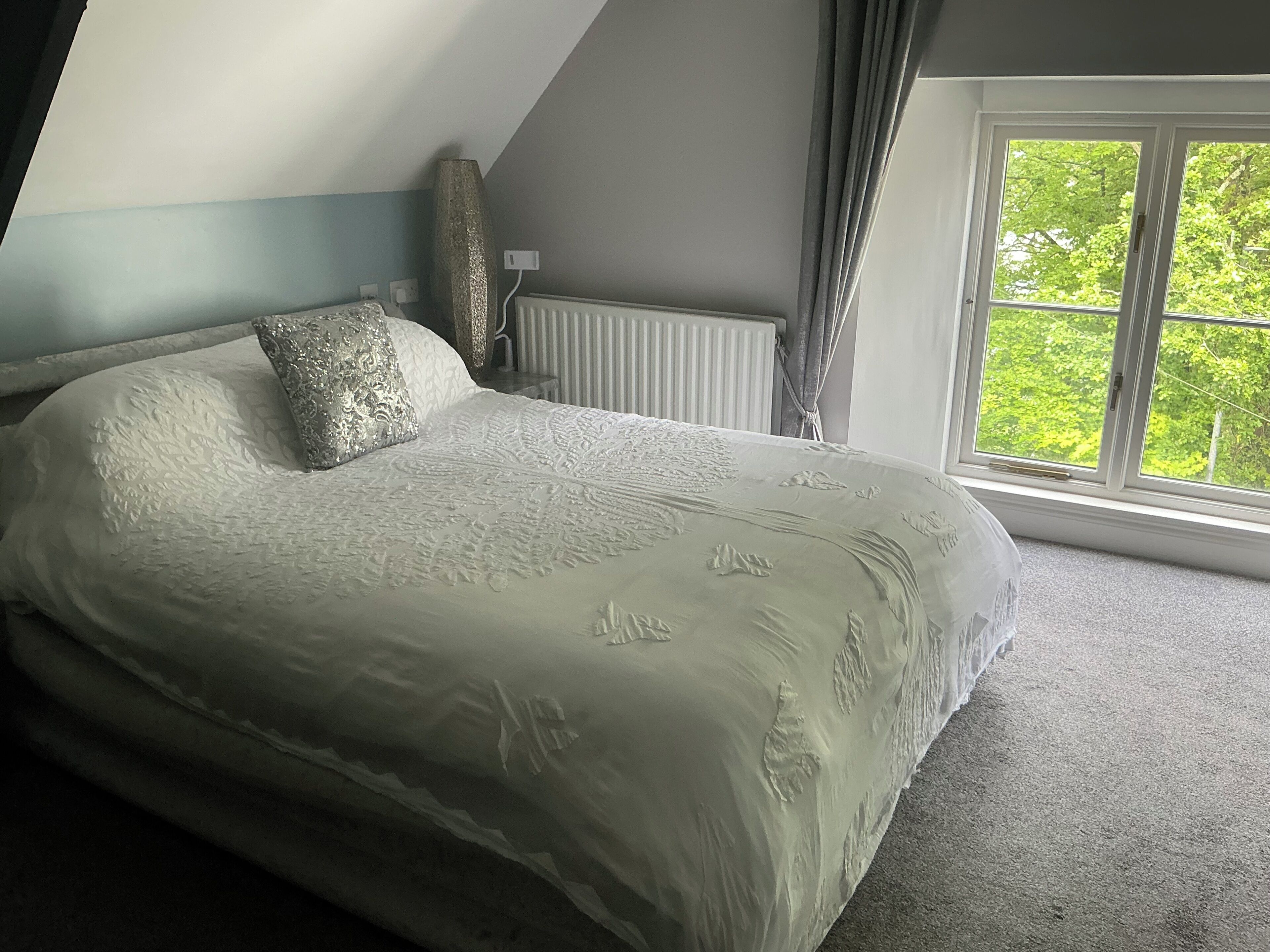A bedroom with a large bed, a radiator, a window with curtains, and a view of greenery outside.