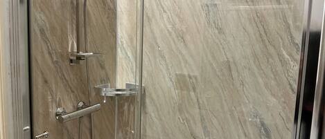 A glass-enclosed shower with a marble wall, a handheld showerhead, and a handheld shower hose.