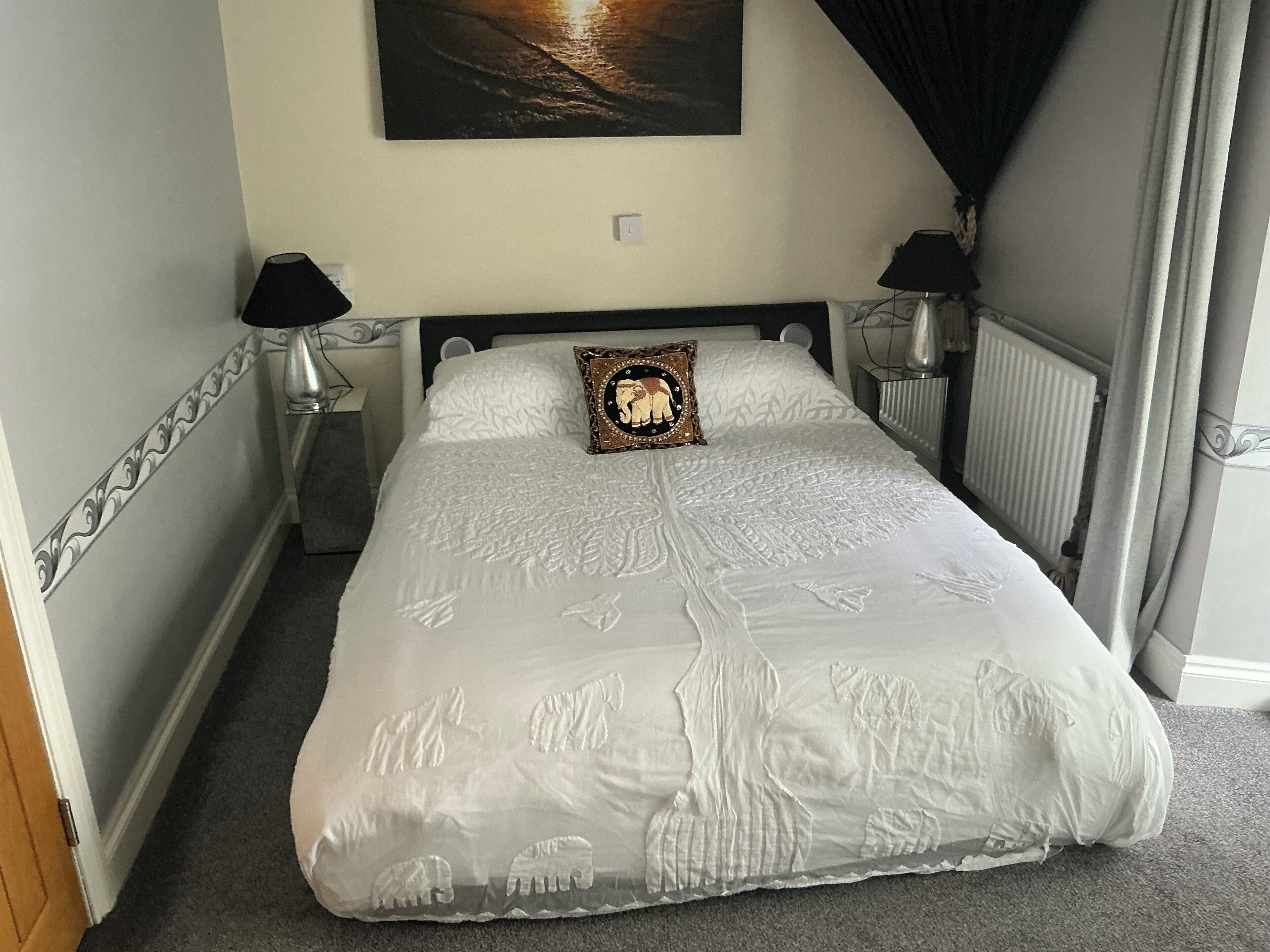A neatly made bed with a white bedspread featuring elephant patterns, two bedside lamps, a wall-mounted picture, and a radiator.