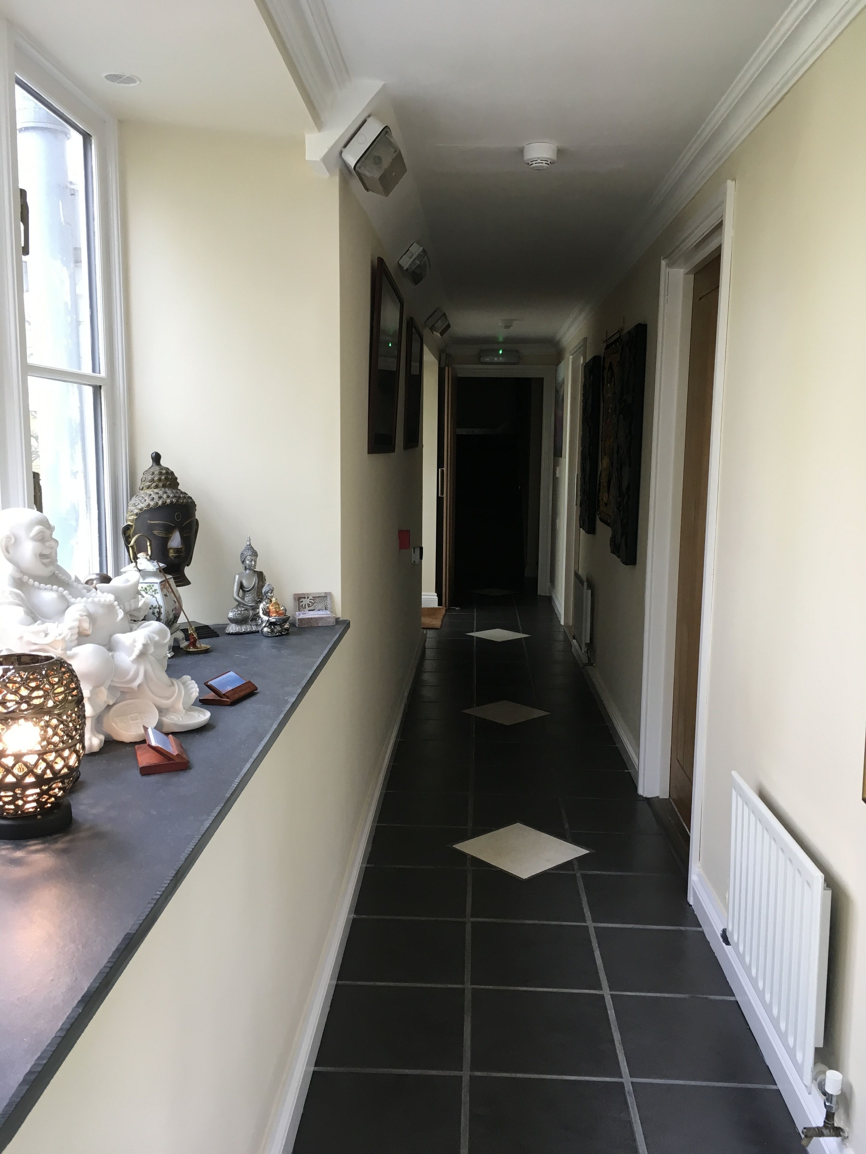 Ein Flur mit schwarzem Fliesenboden, weißen Wänden und einem Fenster links. Statuen und eine brennende Kerze befinden sich ebenfalls auf der linken Seite.
