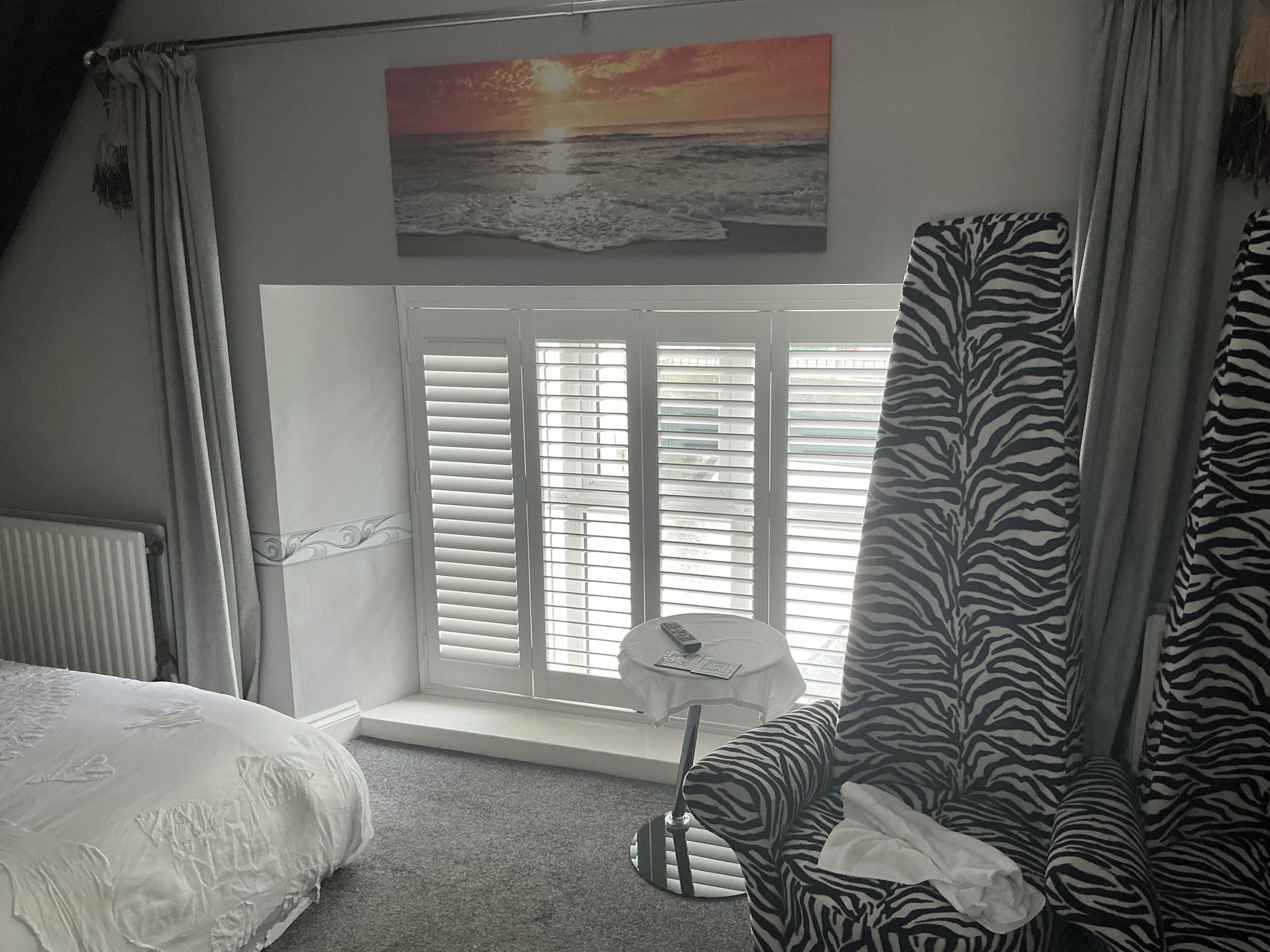 A bedroom with a bed, a zebra-patterned chair, a window with blinds, and a painting of a sunset.