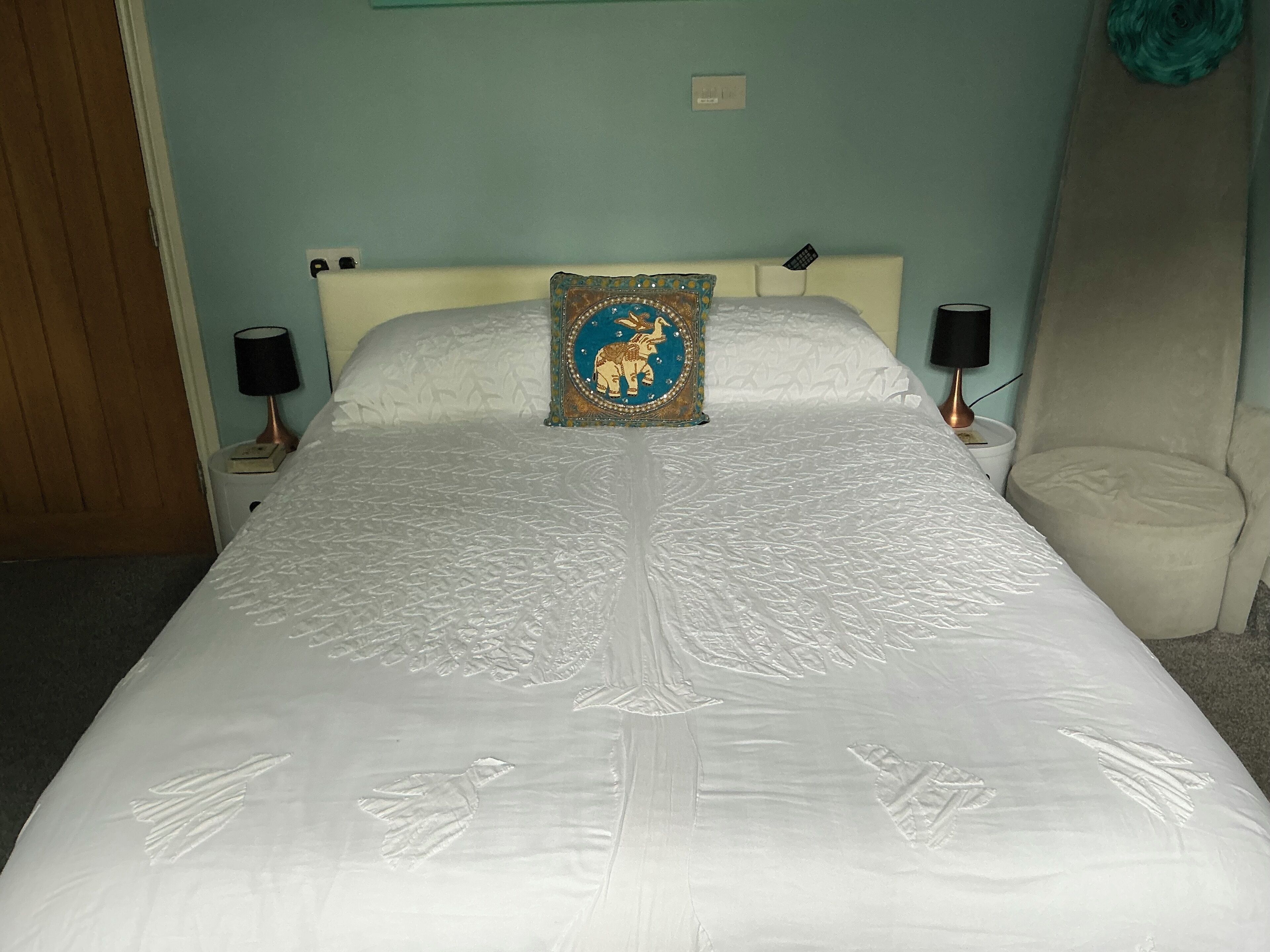 Una cama con colcha blanca, un cojín decorativo, dos lámparas de noche y una silla.