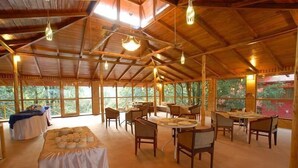 Une vaste salle à manger intérieure, dotée de poutres en bois et de grandes fenêtres. Les tables sont dressées et des chaises sont disposées autour.