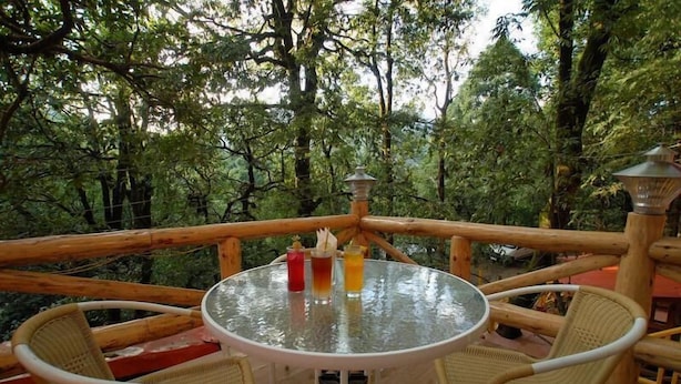 Une table ronde en verre, accompagnée de deux chaises en osier, est placée sur une terrasse en bois dotée d’une rambarde en métal, le tout situé dans un espace boisé.