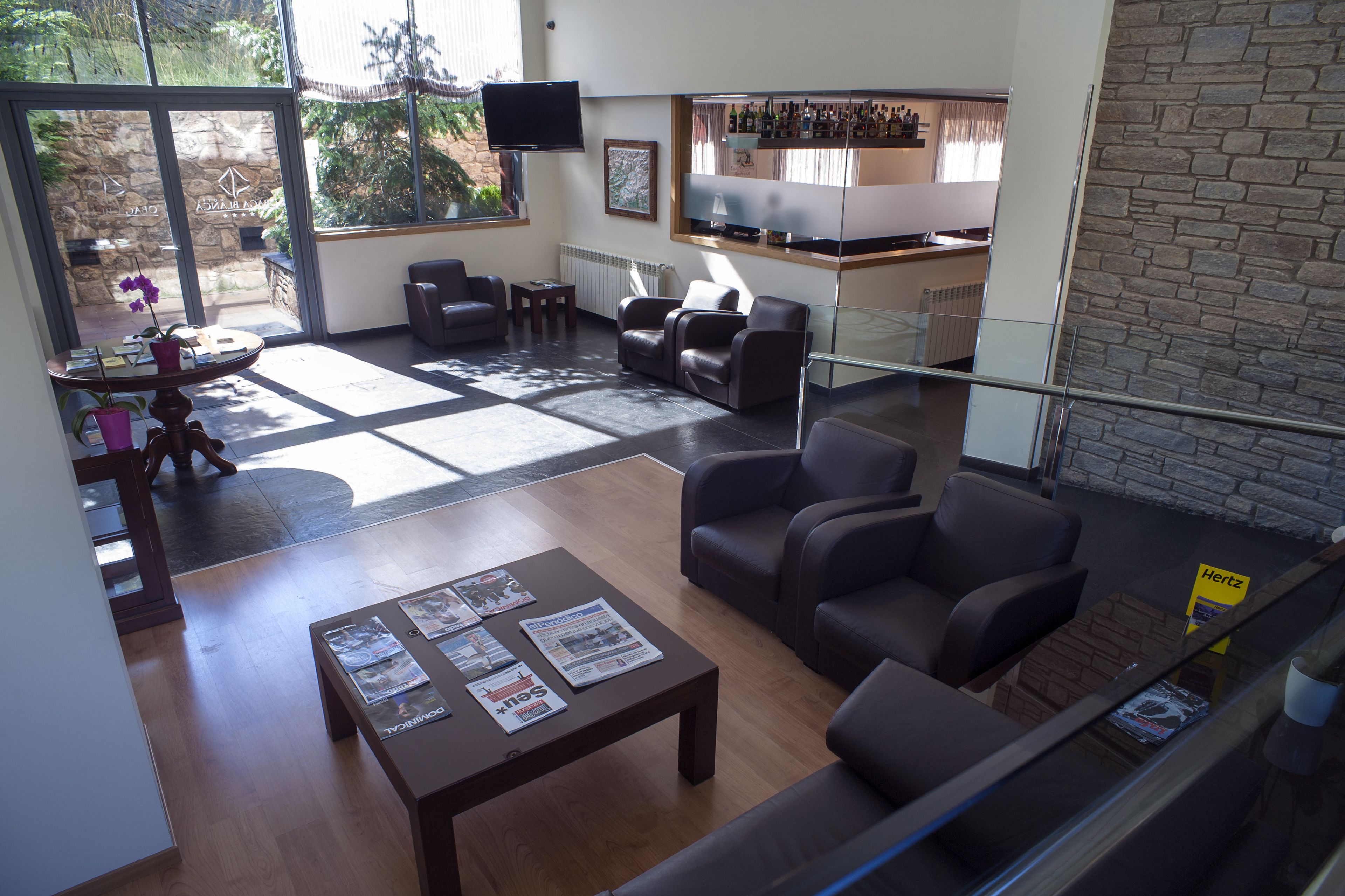 A modern living room with leather sofas, a coffee table with magazines, a stone wall, and a kitchen area with a bar.