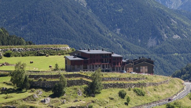 A large, multi-story building with a stone foundation and a flat roof, surrounded by a lush green landscape and mountains in the background. - Hotel Obaga Blanca (Canillo)