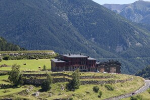A large, multi-story building with a stone foundation and a flat roof, surrounded by a lush green landscape and mountains in the background.