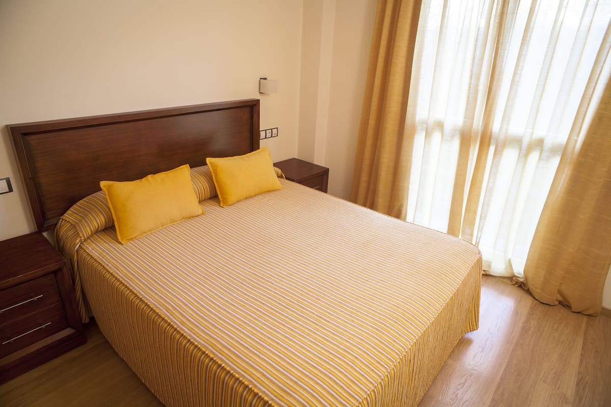 A neatly made bed with yellow pillows and striped bedding, a wooden headboard, a bedside table, and a window with curtains.