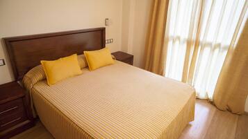 A neatly made bed with yellow pillows and striped bedding, a wooden headboard, a bedside table, and a window with curtains.