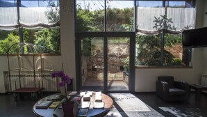 A room with a table displaying books and a potted plant, a chair, and a view through a glass door to an outdoor area with a stone wall and greenery. - Hotel Obaga Blanca (Canillo)