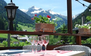 Una mesa con dos copas de vino y un tenedor, con vistas a montañas, una baranda de madera y macetas con flores.
