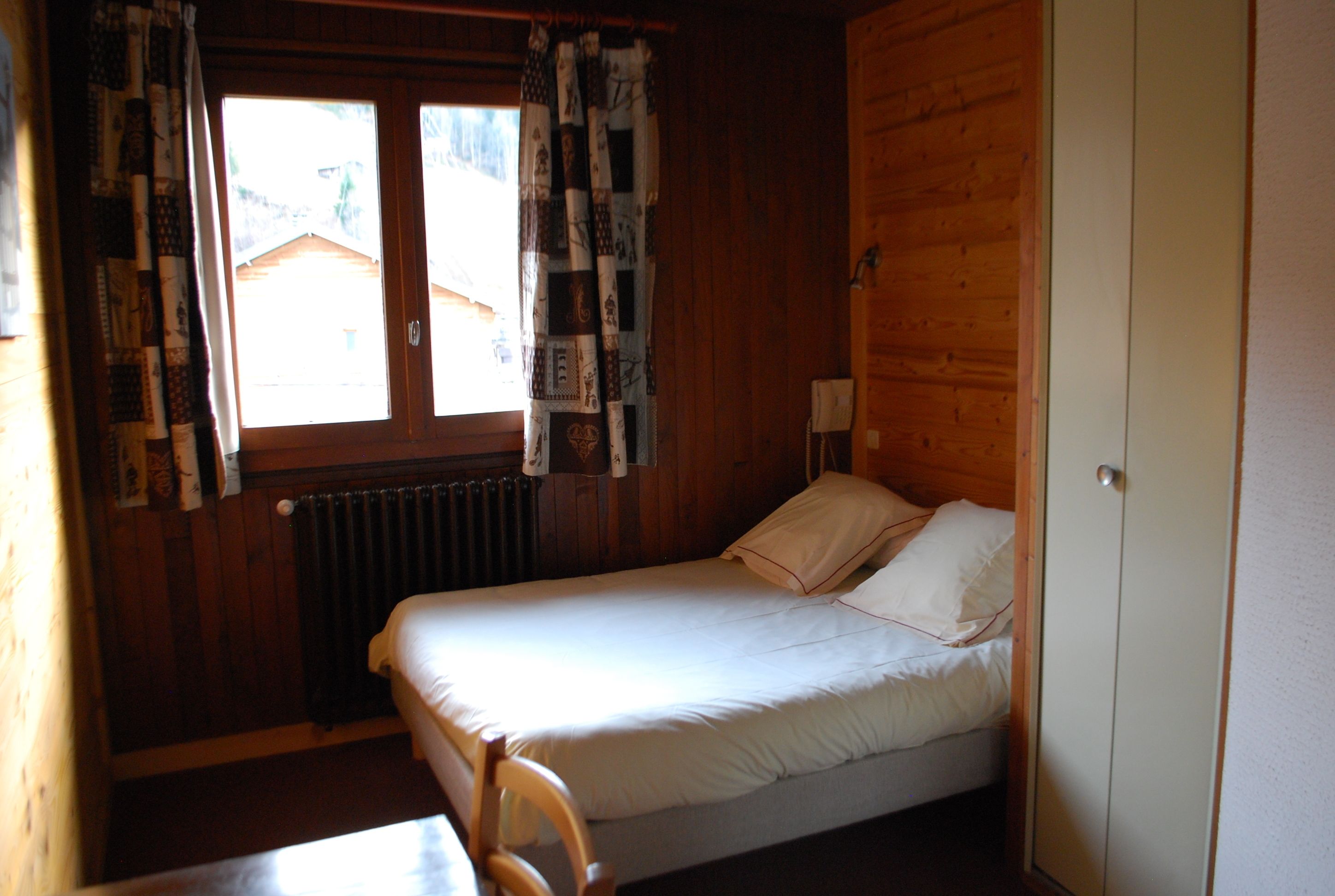Una stanza piccola con un letto singolo, pavimento in legno, una finestra con tende, un termosifone e una porta.