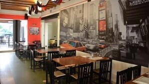 Interior de un restaurante con mesas y sillas de madera, una pared de acento roja y un mural grande que representa una calle de la ciudad.