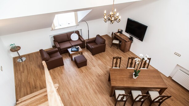 A modern loft apartment with a wooden floor, a dining area with a wooden table and chairs, a sofa set, a TV mounted on the wall, and a staircase.
