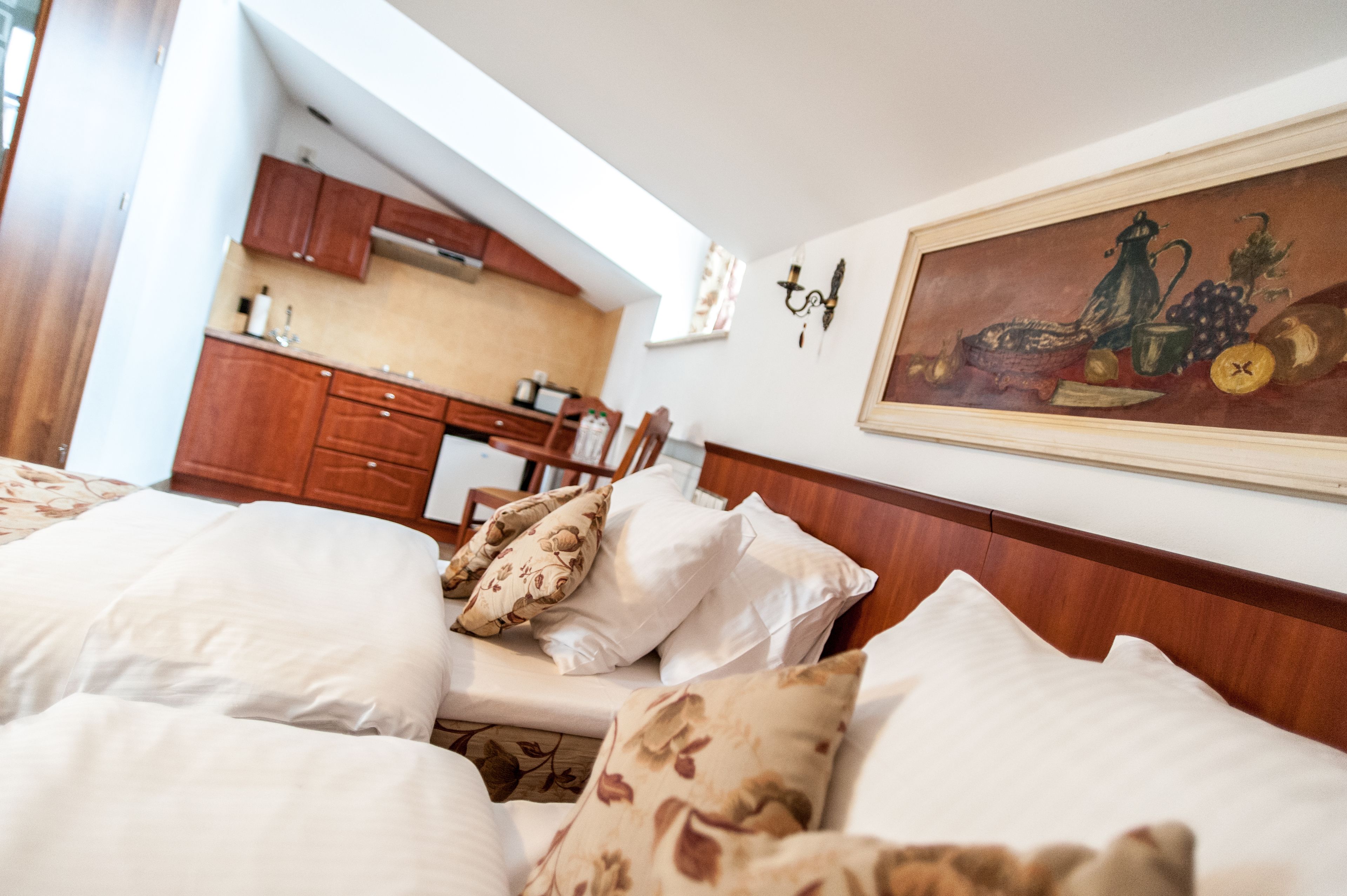 A bedroom with a large bed, a painting on the wall, and a view of a kitchen in the background.