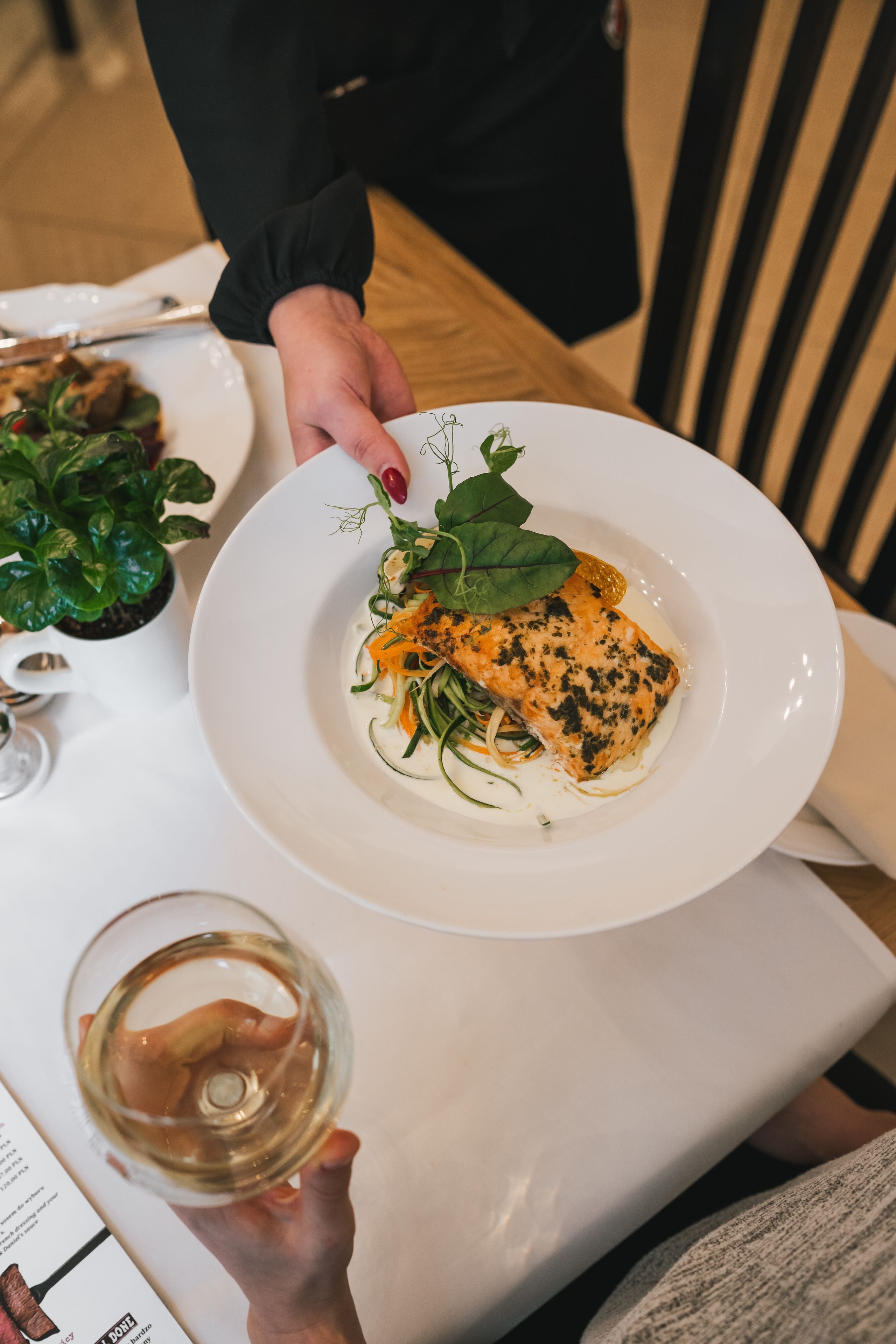 A person holding a white plate with a salmon dish, a glass of white wine, and a menu.