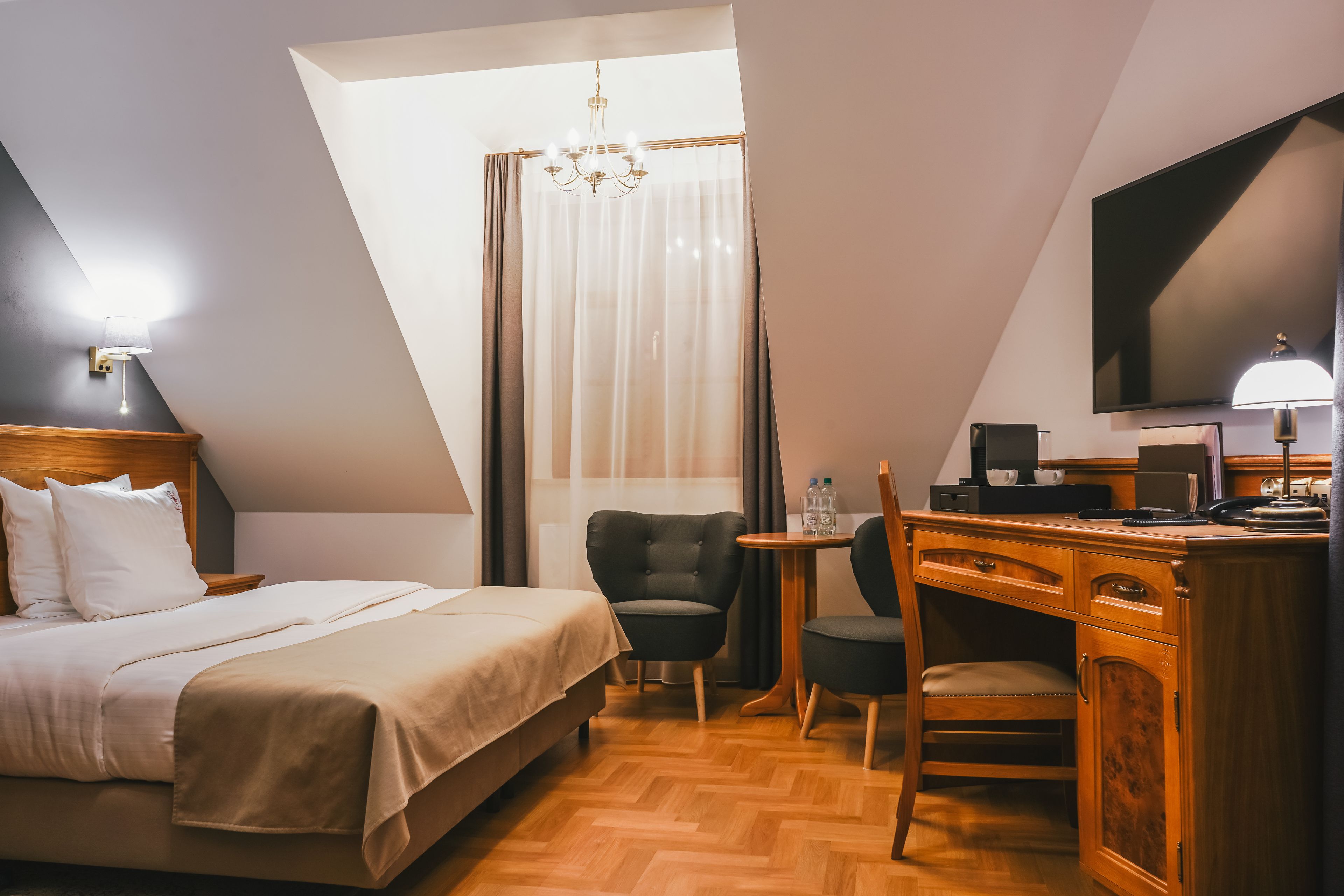Ett hotellrum med en säng, ett skrivbord, stolar, en tv och en kristallkrona.