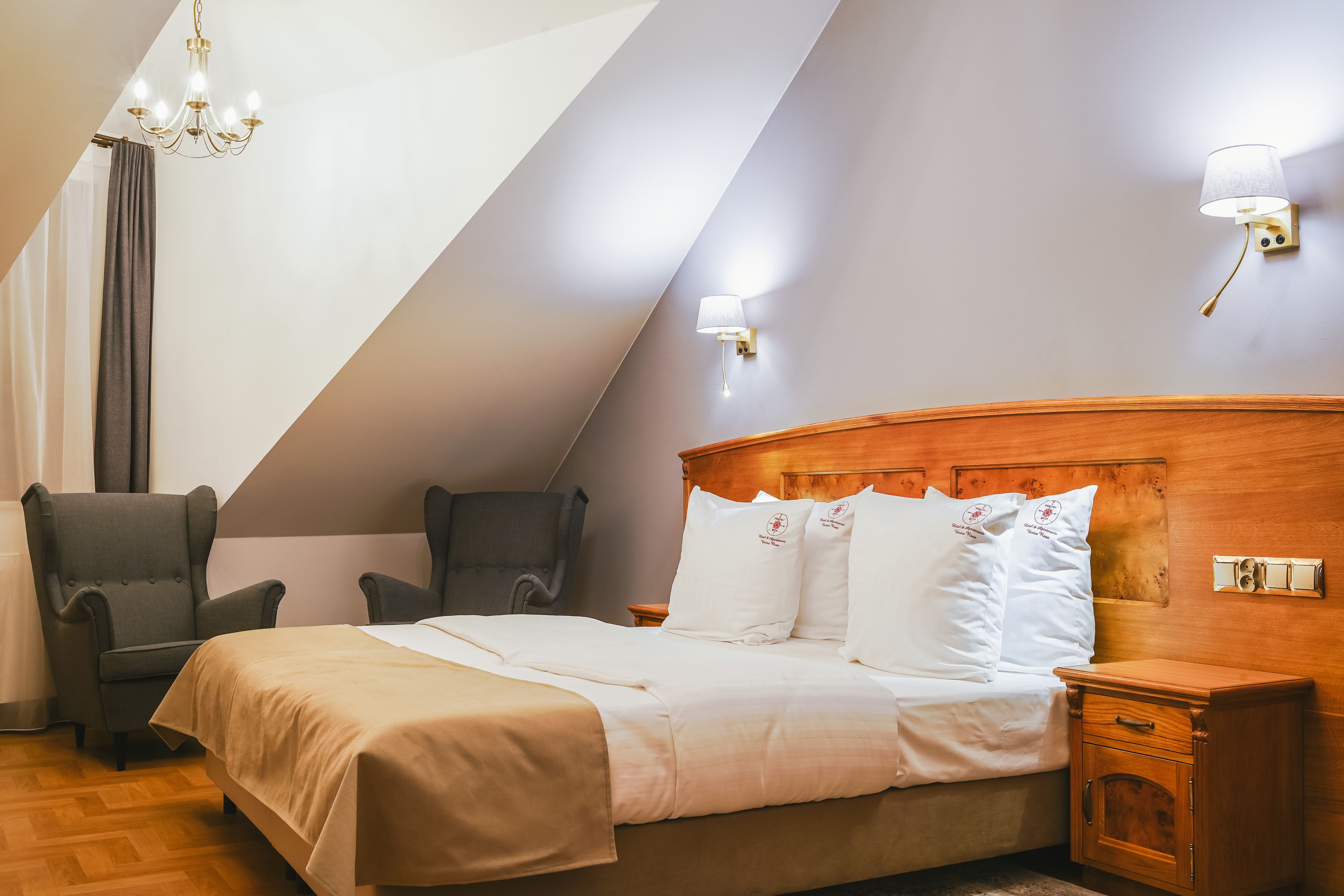Een slaapkamer met een bed, twee fauteuils, een nachtkastje, een houten nachttafel, een kroonluchter en een wandlamp.