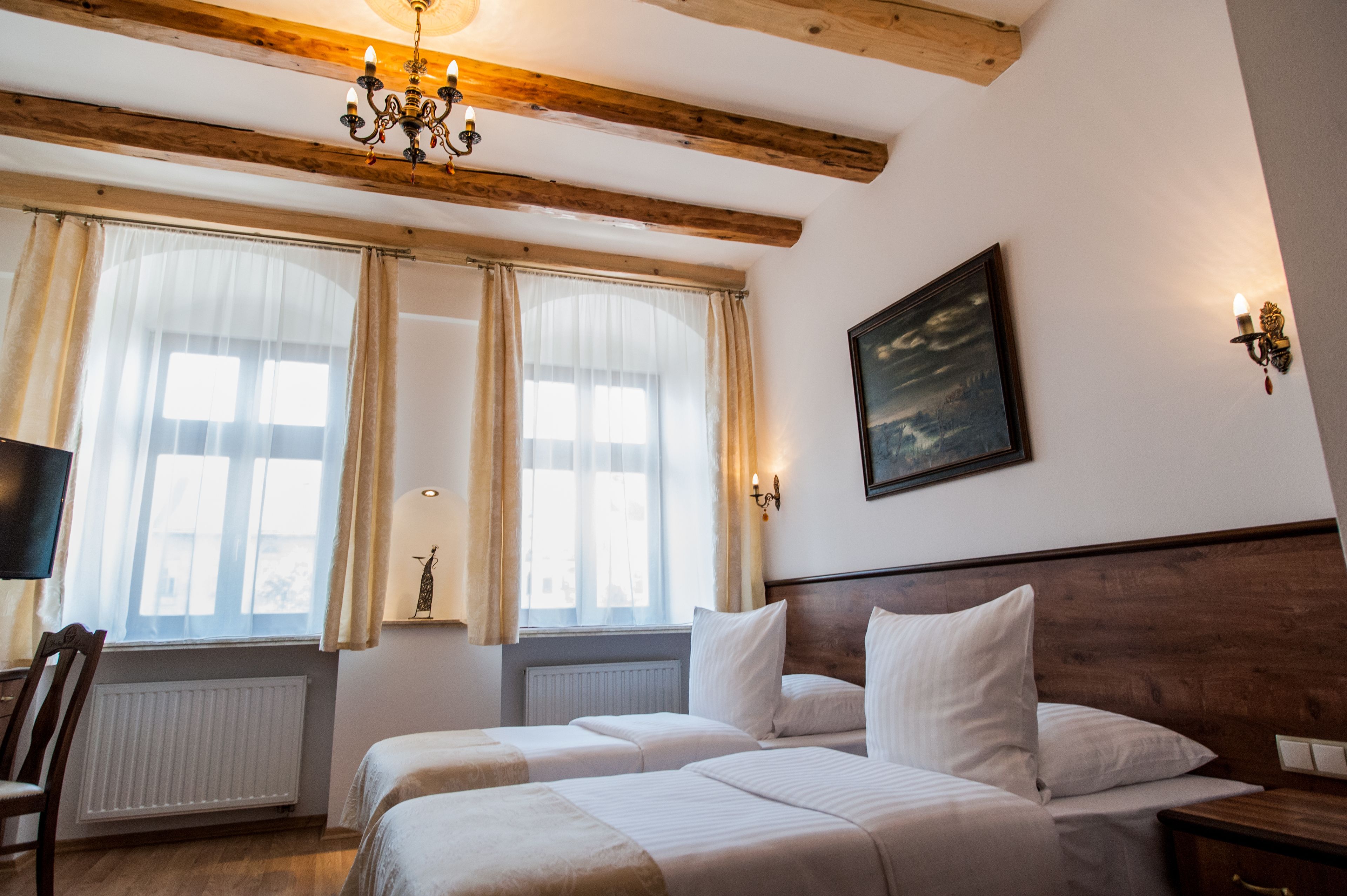 A hotel room with two beds, a wooden ceiling, a chandelier, a painting on the wall, and a window with curtains.