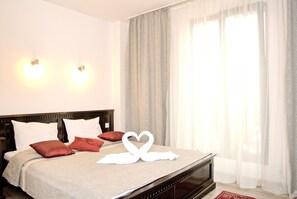 A neatly made bed with a heart-shaped towel arrangement, a wooden headboard, and a view of a balcony through sheer curtains. - Belleville (Iasi)
