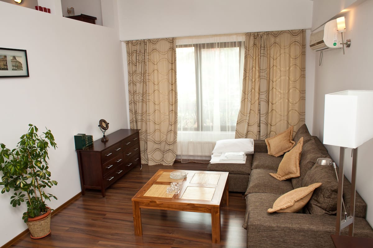 A living room with a wooden coffee table, a sofa, a potted plant, a dresser, and a window with curtains.
