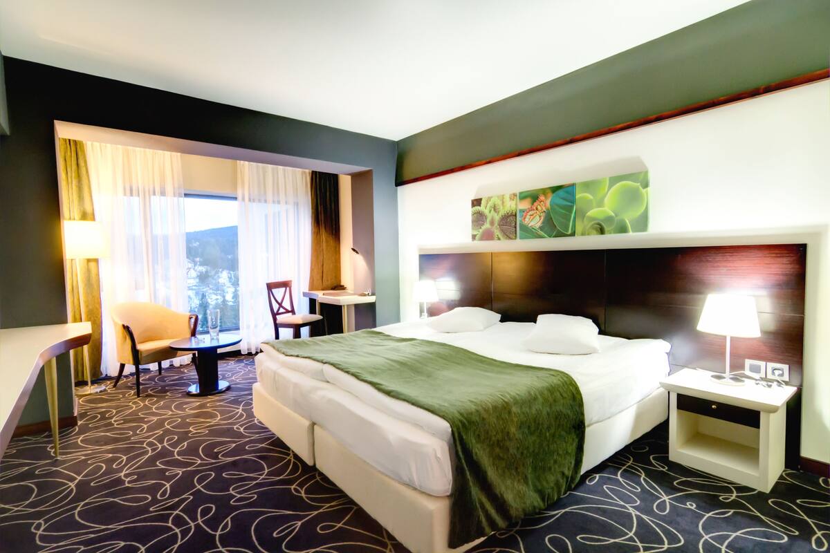 Ein Hotelzimmer mit einem großen Bett, einem Schreibtisch, einem Sessel und Blick ins Freie.