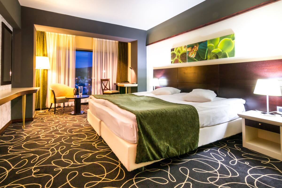 Ein Hotelzimmer mit einem großen Bett, einem Schreibtisch, einem Stuhl und Blick auf die beleuchtete Stadt.