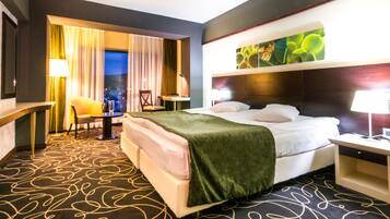 A hotel room with a large bed, a desk, a chair, and a view of the city at night.