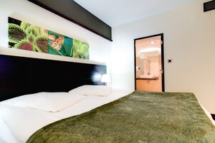 Una habitación de hotel moderna con una cama grande, cabecera, una obra de arte colgada en la pared, una puerta y un baño visible a través de una puerta abierta.
