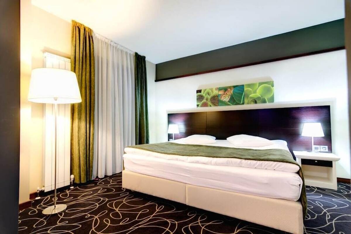 Ein Hotelzimmer mit einem Bett, einem Nachttisch, einer Lampe, einem Bild an der Wand und einem Fenster mit Vorhängen.