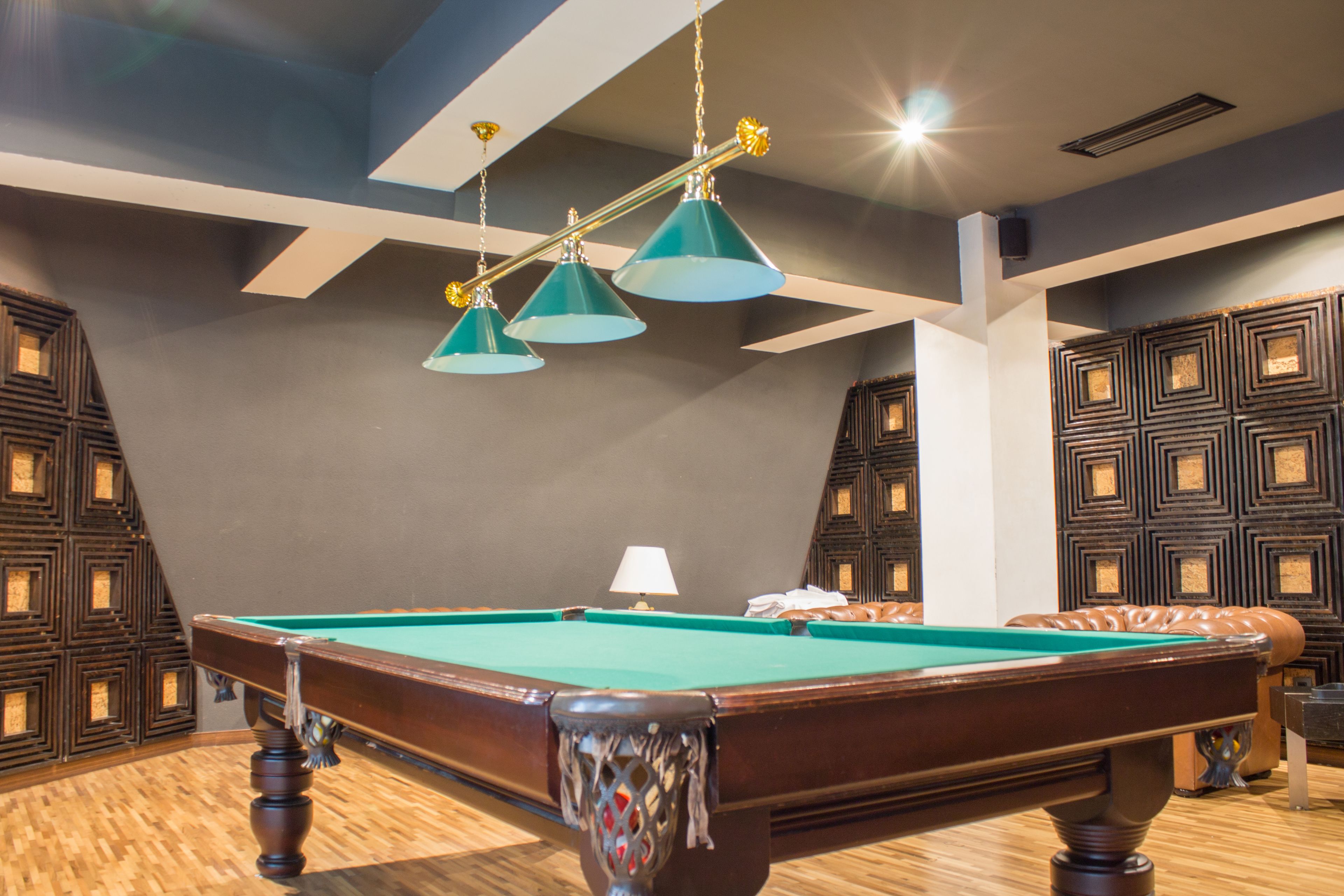 Een pooltafel met een groen vilt oppervlak, omgeven door houten vloeren. Drie hanglampen met blauwe kap en gouden accenten. De kamer heeft een modern design met geometrische wandpatronen en een bruine leren bank.
