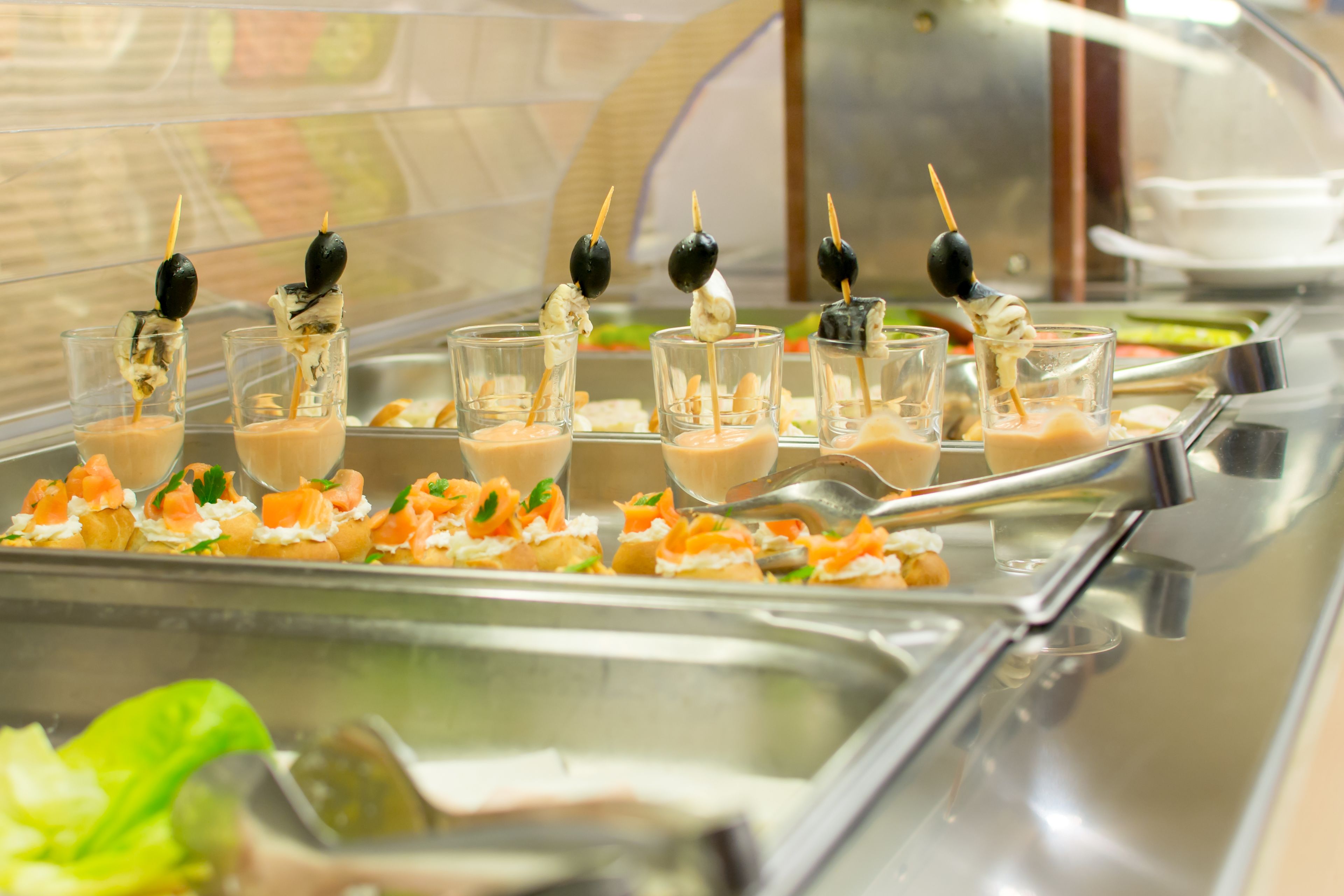 Un bufé con diferentes opciones de comida, incluyendo pequeñas copas con una sustancia cremosa y adornadas con hierbas, y brochetas con aceitunas y otros adornos.