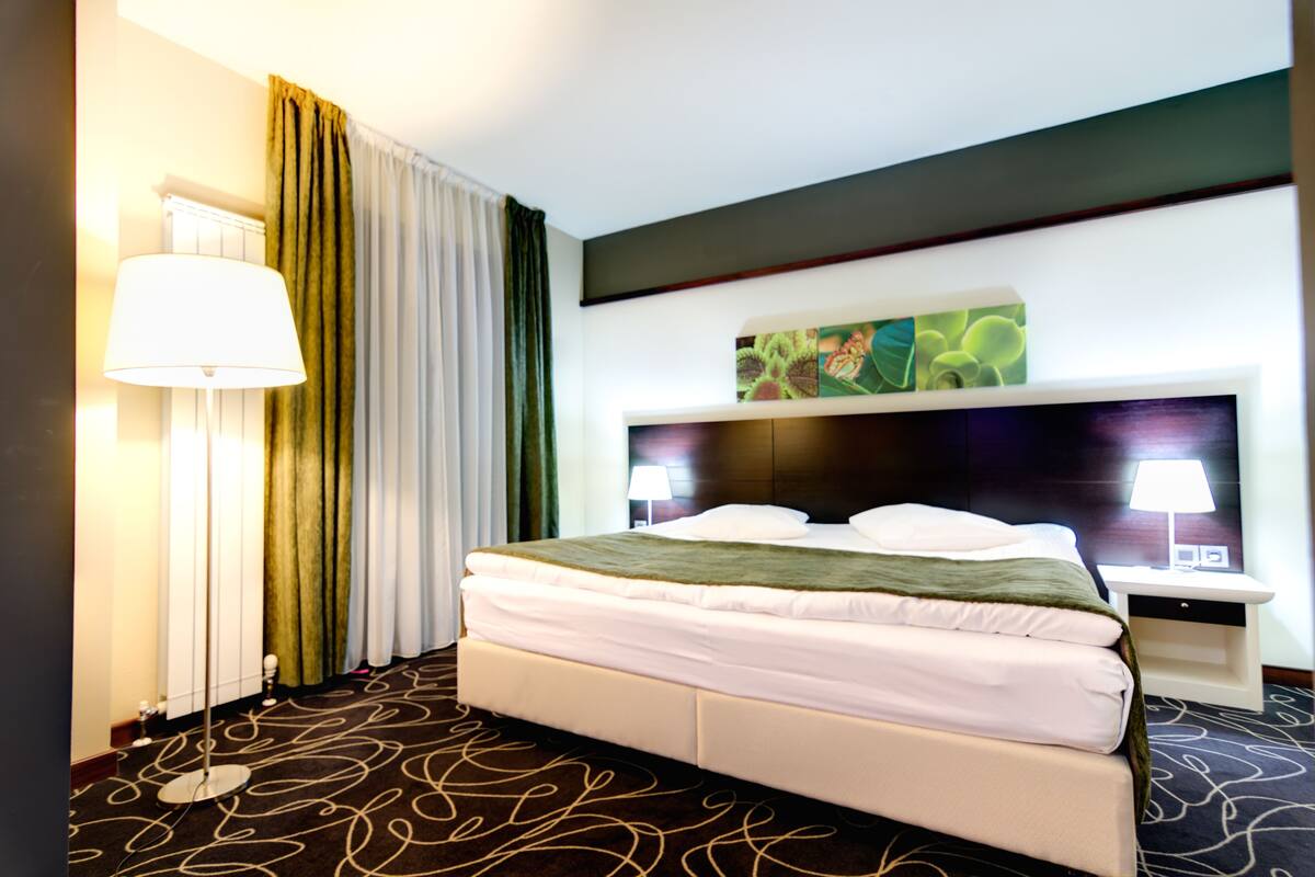 Ein Hotelzimmer mit einem großen Bett, einem Nachttisch mit Lampe, einem an der Wand montierten Fernseher und einer Zierpflanze.