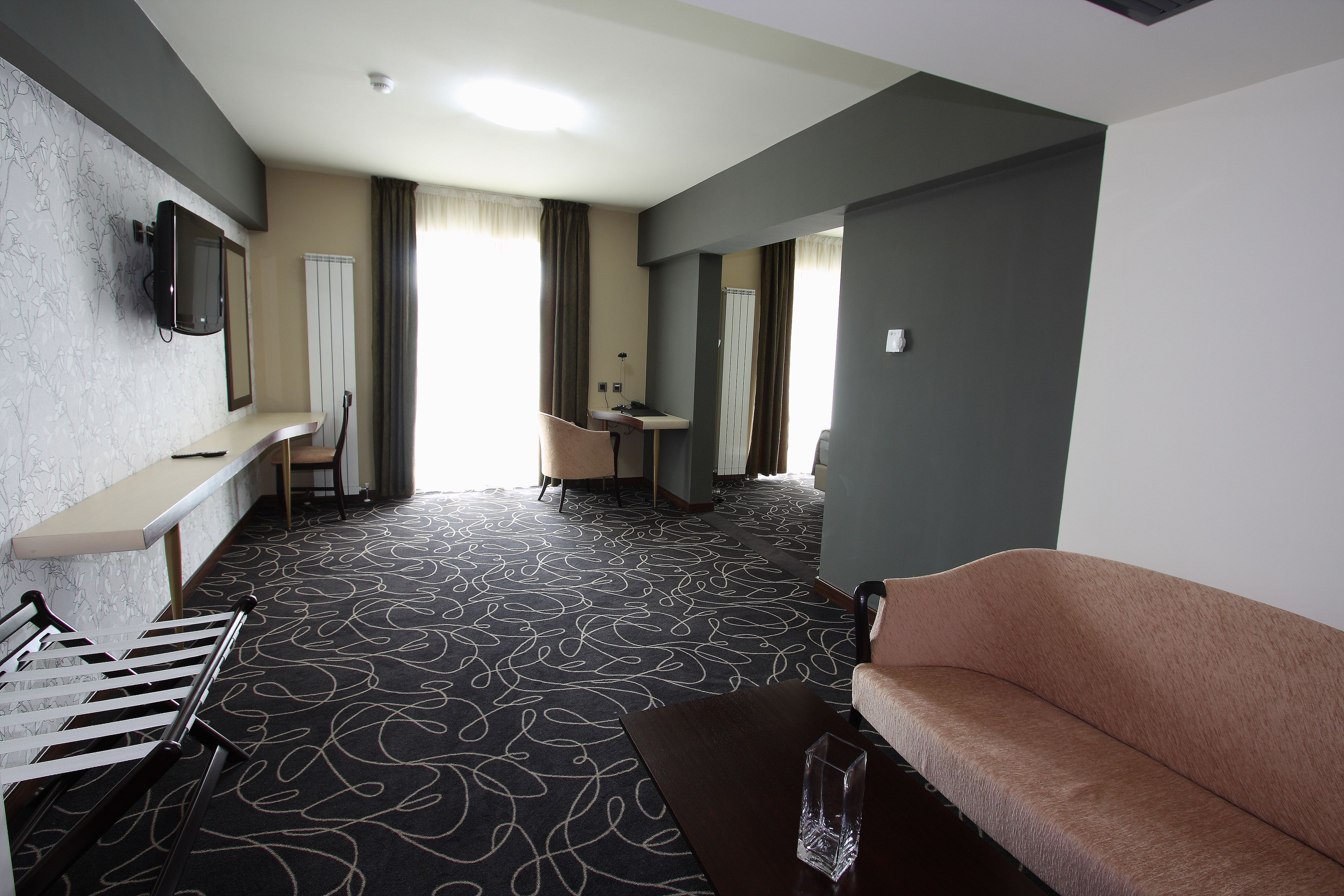 A hotel room with a sofa, a small table, a TV mounted on the wall, a desk with a chair, and a patterned carpet.