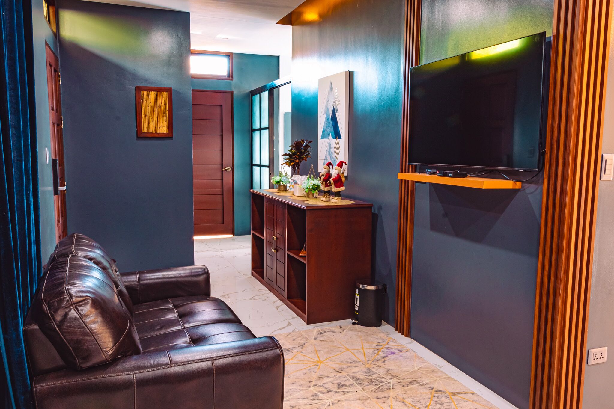 Un salon moderne avec un canapé en cuir, un téléviseur fixé sur un panneau en bois et un petit meuble en bois orné d’objets décoratifs.